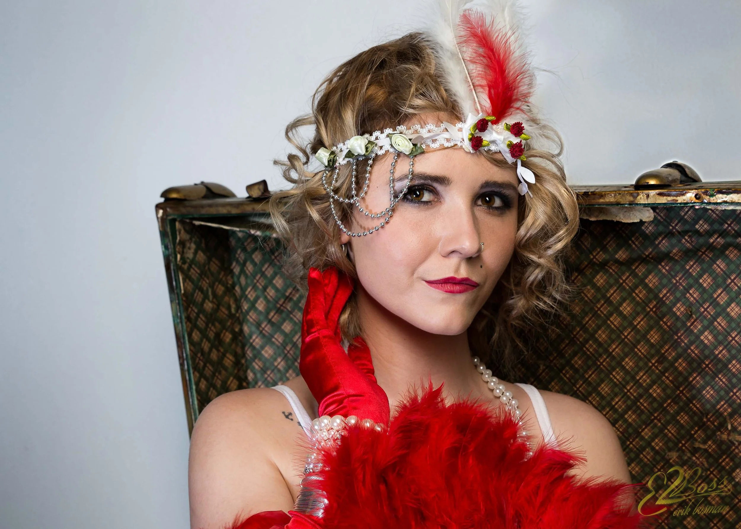 Woman with curly blonde hair wearing a flowered headband with a red feather, red gloves, pearl necklace, and makeup, holding a red feathered fan, standing in front of a vintage suitcase.