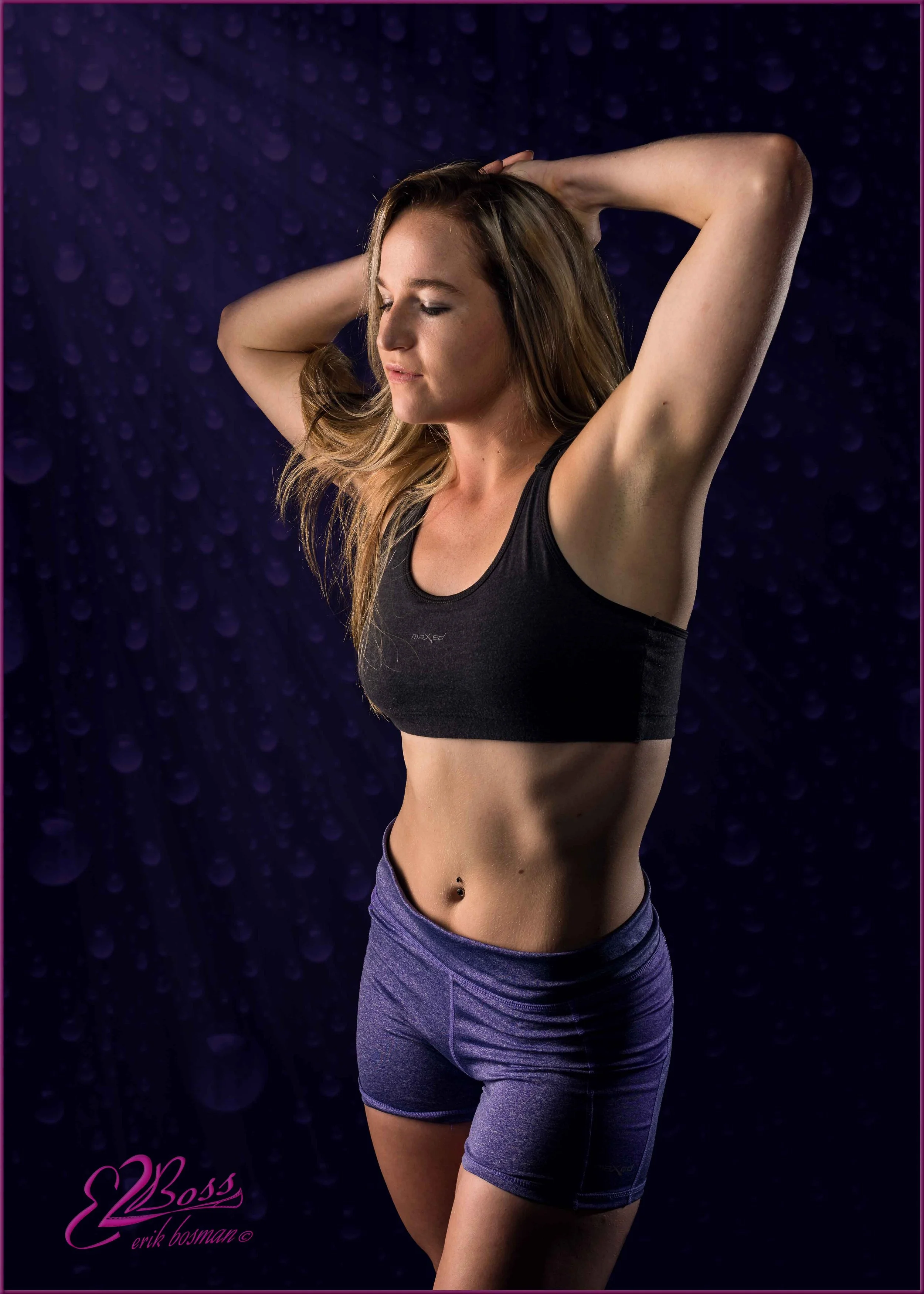 A woman with long blond hair wearing a black sports bra and purple shorts, posing with her eyes closed and arms raised behind her head against a dark purple background with bokeh effect.