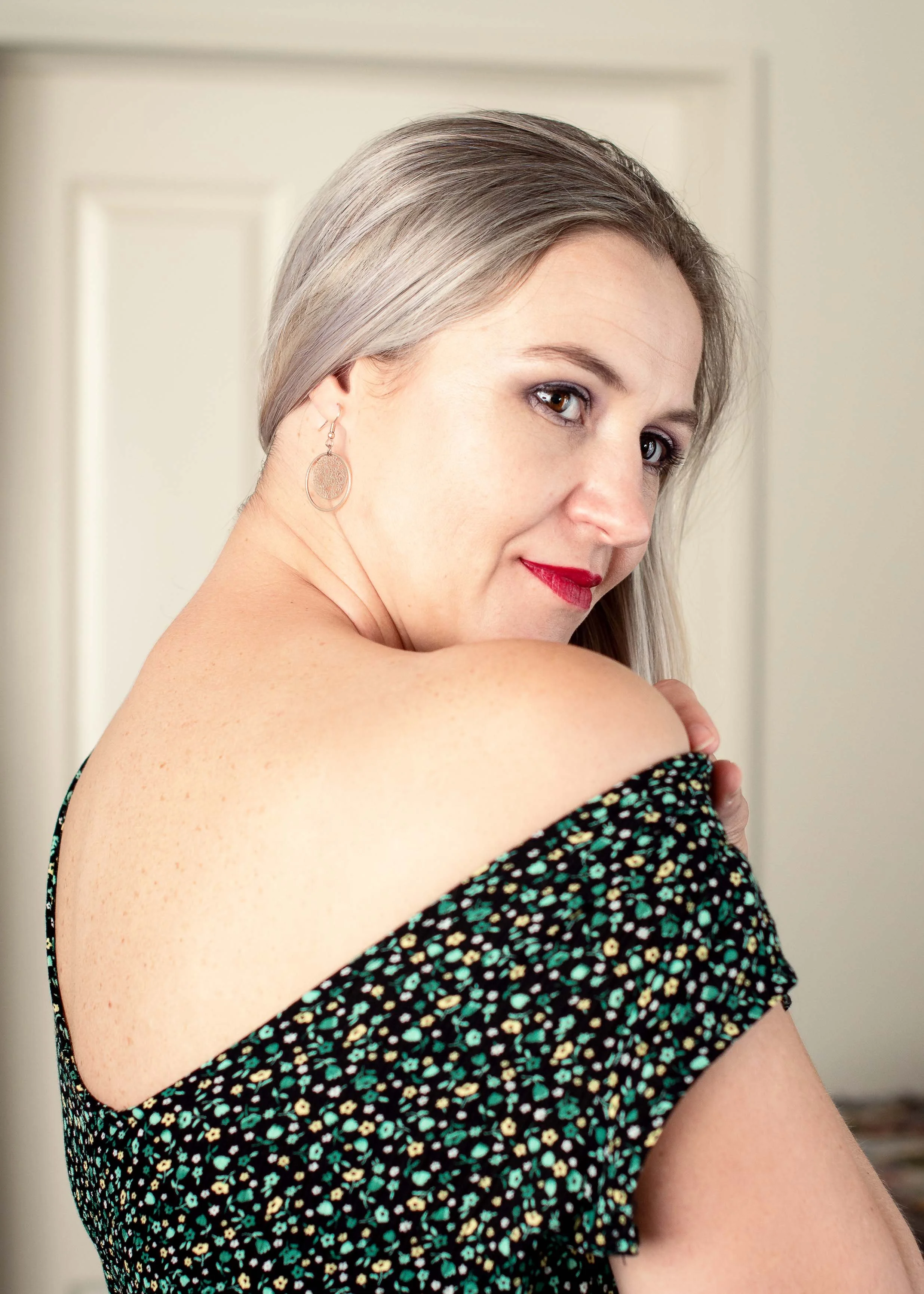 A woman with silver hair and red lipstick poses with her shoulder exposed, wearing a black dress with a colorful floral pattern and round earrings.