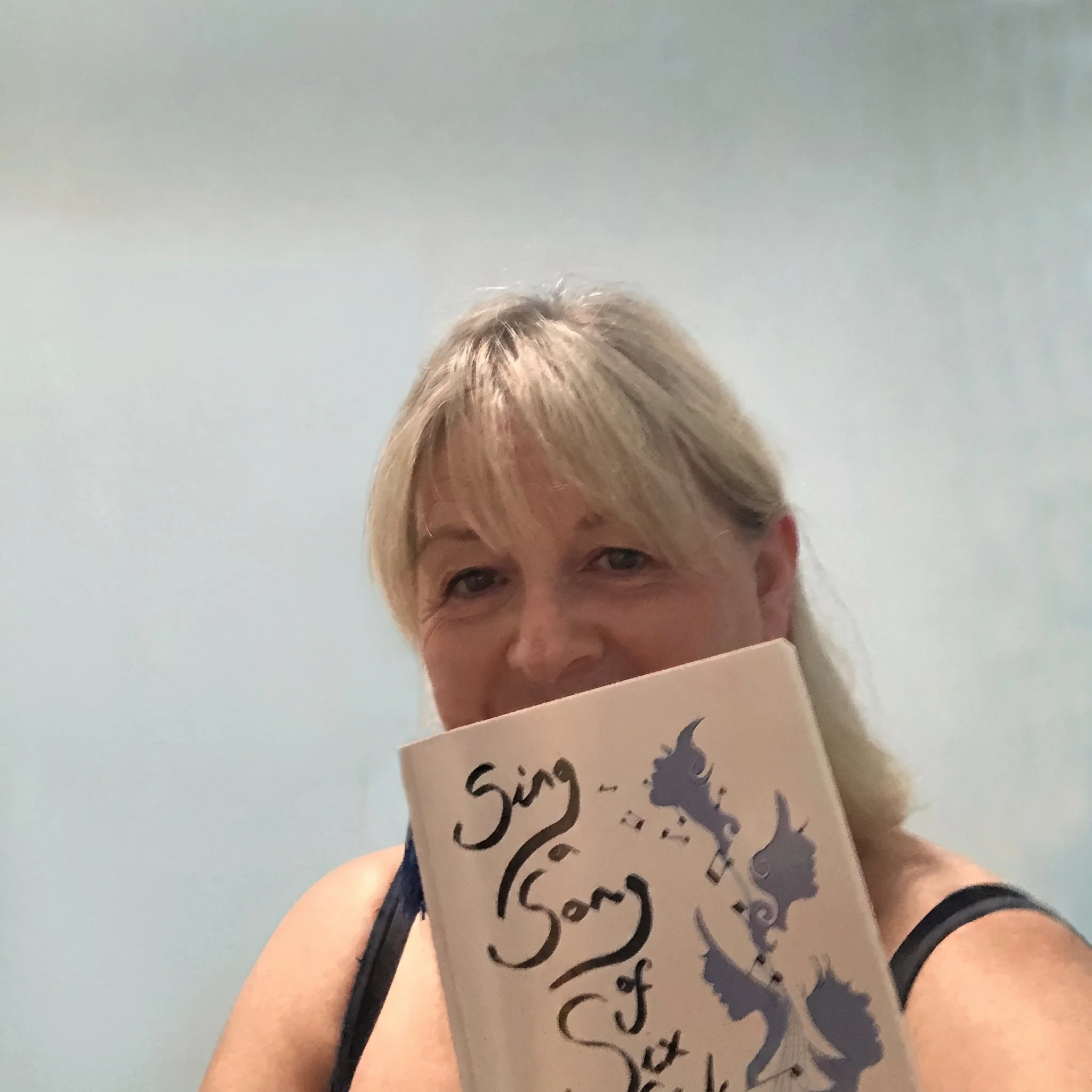 A person with short blonde hair holding a book entitled "Sing a Song of Six Girls!" and some other text in parentheses, with part of their face visible and a light blue background.