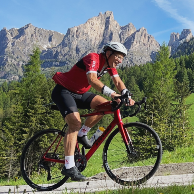 Cyclist rides on road with mountains in background, wearing red jersey and helmet.