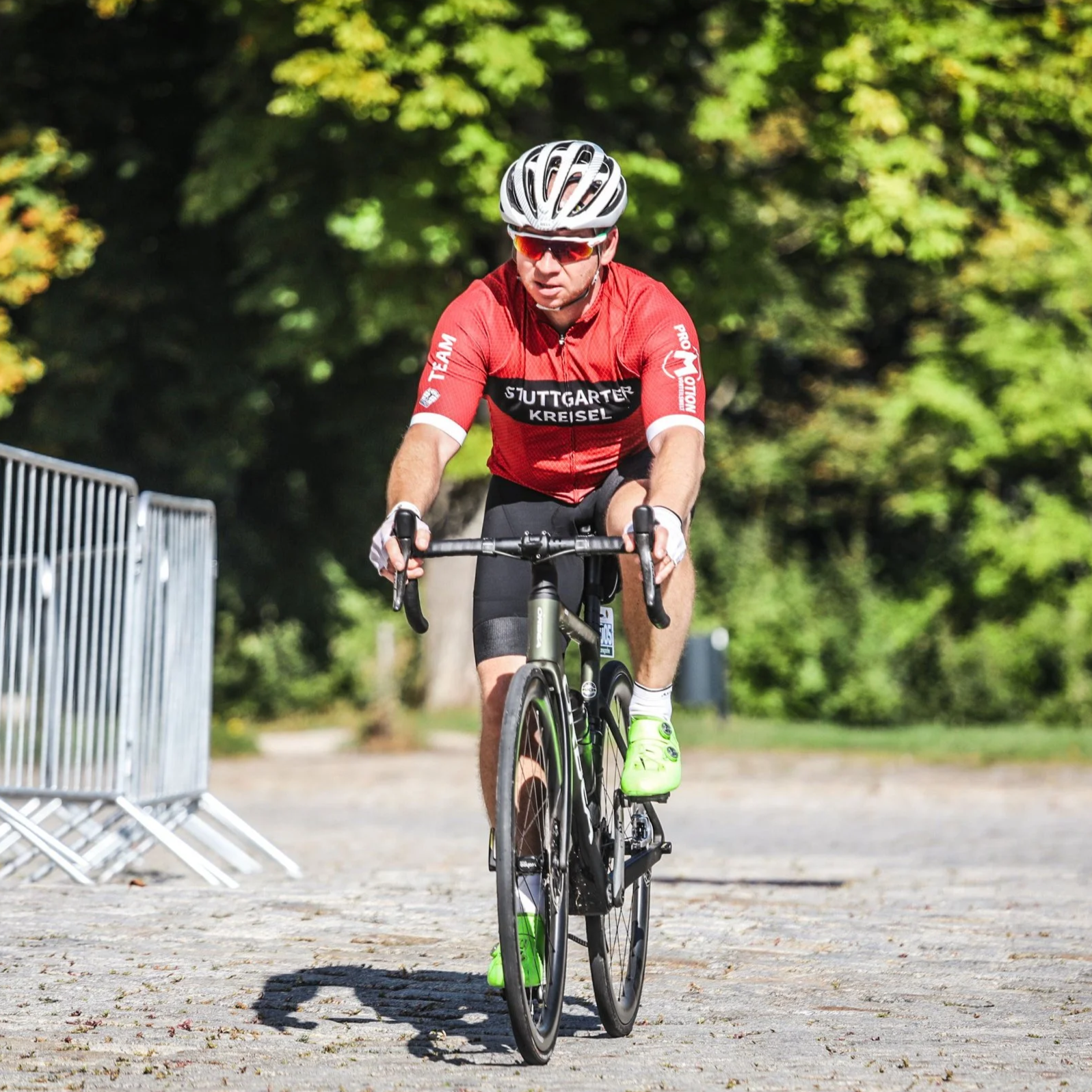 Cyclist wearing red jersey, helmet, and sunglasses riding on cobblestone path with metal barriers and trees in background.