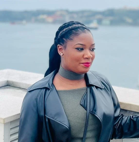 Jeune femme avec des cheveux tressés, portant une veste en cuir noir, un haut gris et un collier ras du cou, se tenant près d'une rambarde avec un plan d'eau en arrière-plan.