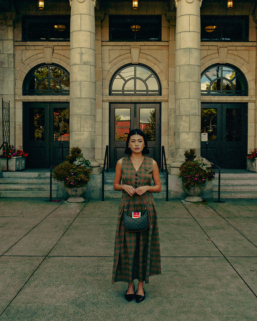 A woman standing outside a grand stone building with large arched windows and columns, wearing a sleeveless plaid dress, carrying a handbag, and looking at the camera.