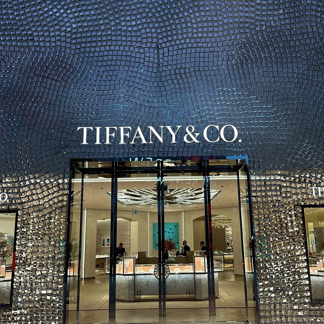 Exterior view of the Tiffany & Co. store entrance with a mosaic tiled wall and glass doors, showcasing the interior with staff and display cases.