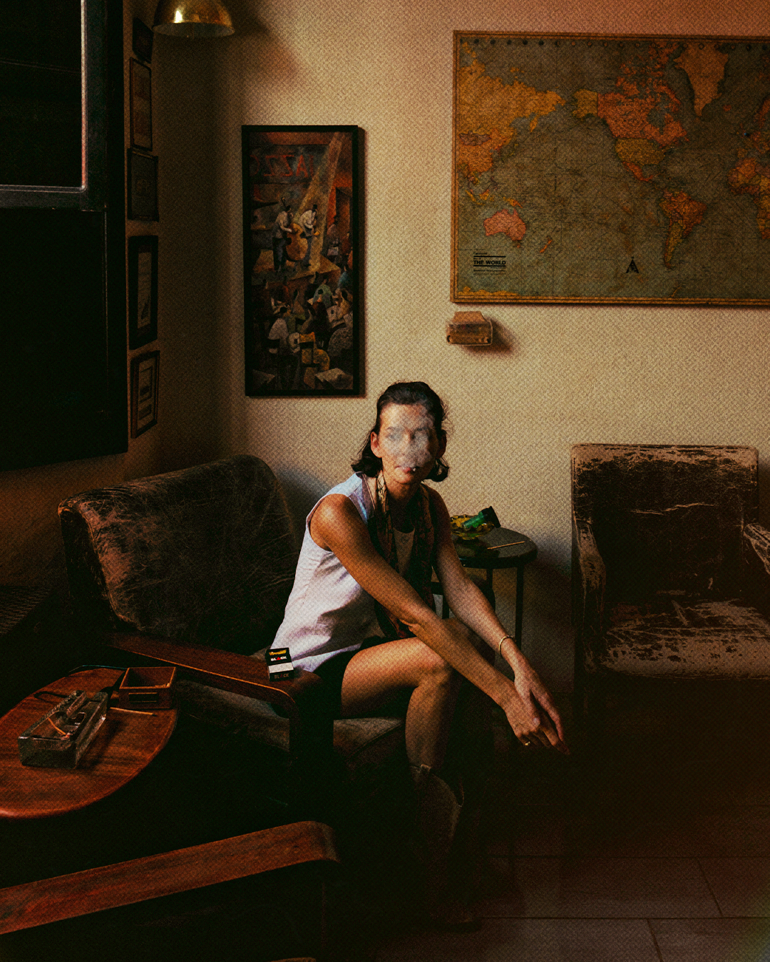Woman sitting on a vintage couch smoking with a cloud of smoke visible, in a cozy, dimly lit room decorated with world maps and framed art on the walls.