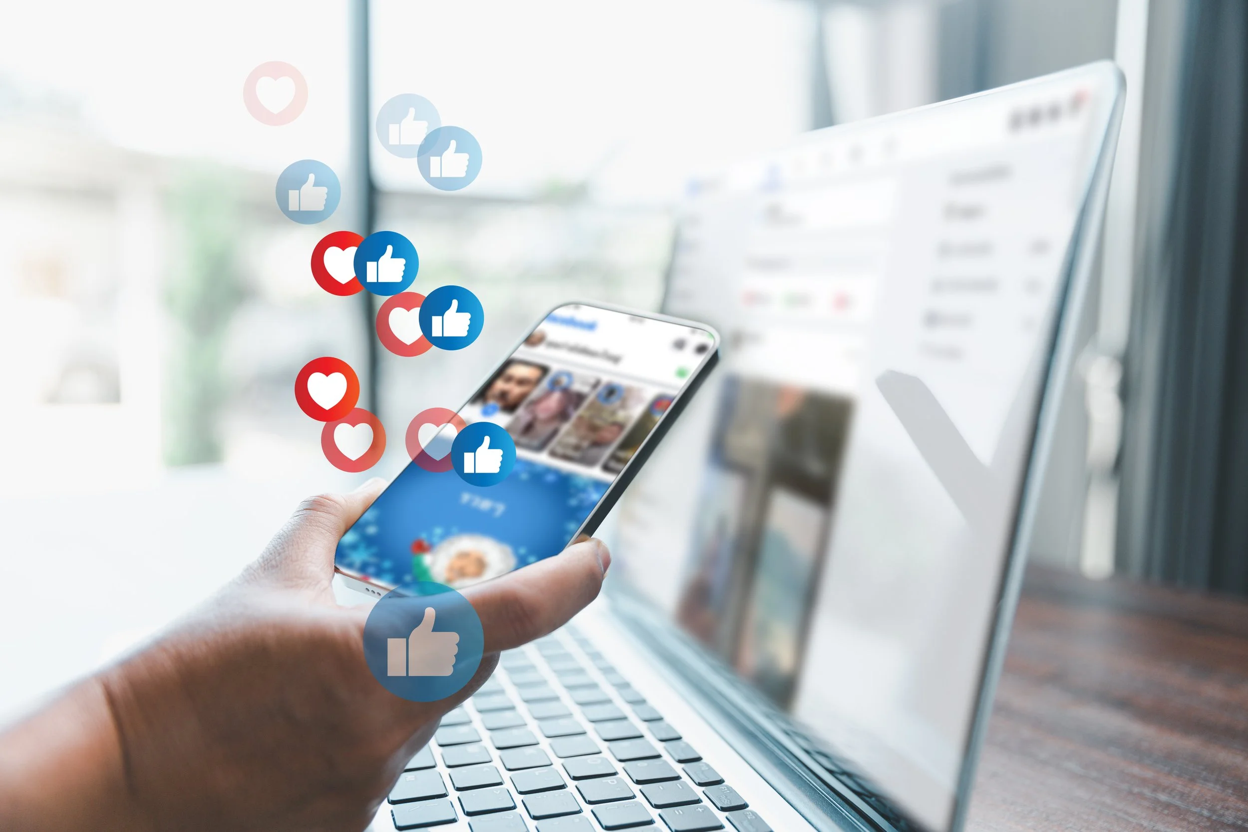 Person holding a smartphone with Facebook likes and hearts floating above, in front of a laptop on a desk.