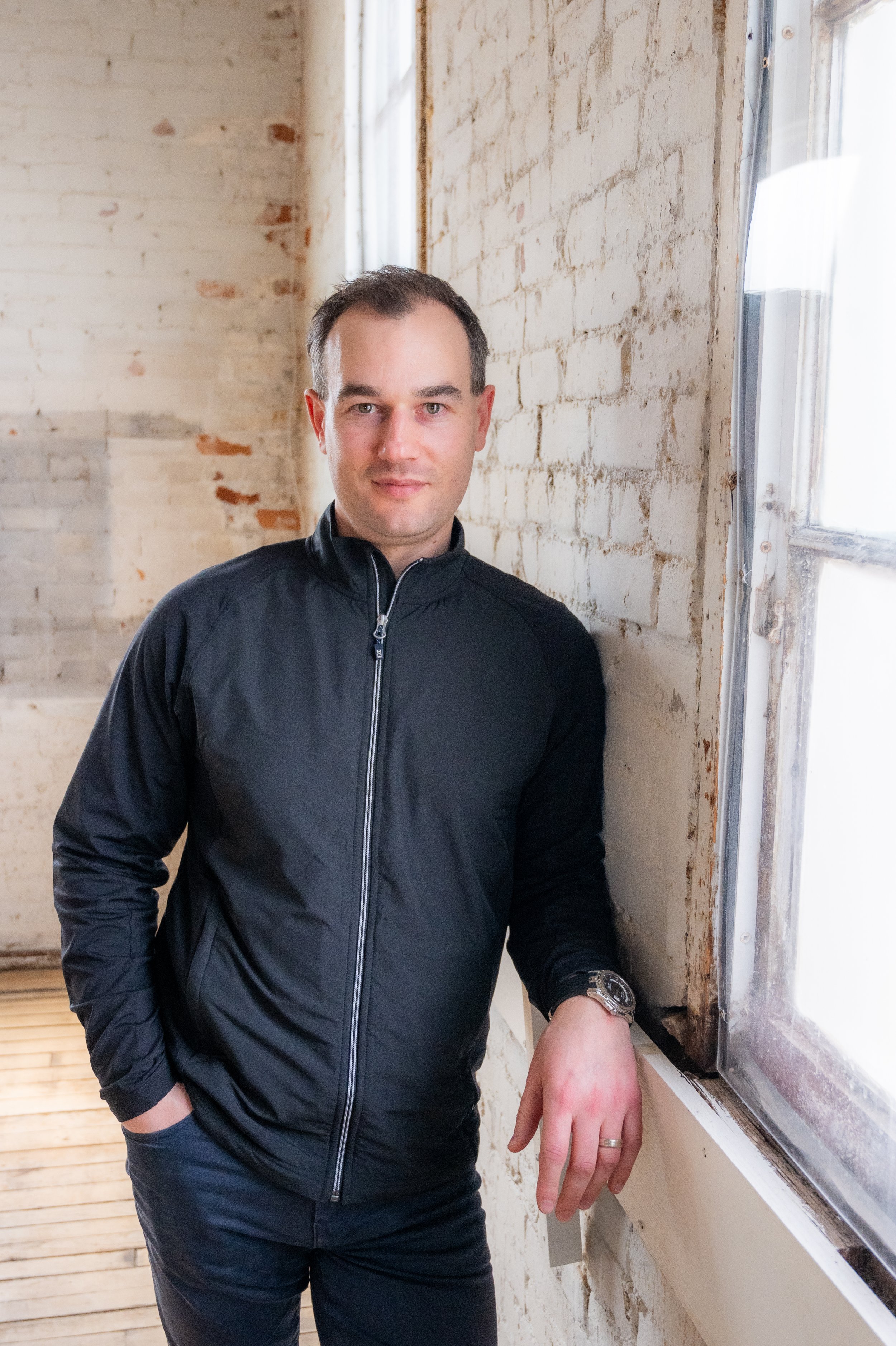 Man in black zip-up jacket standing indoors near a window with white brick wall in the background.