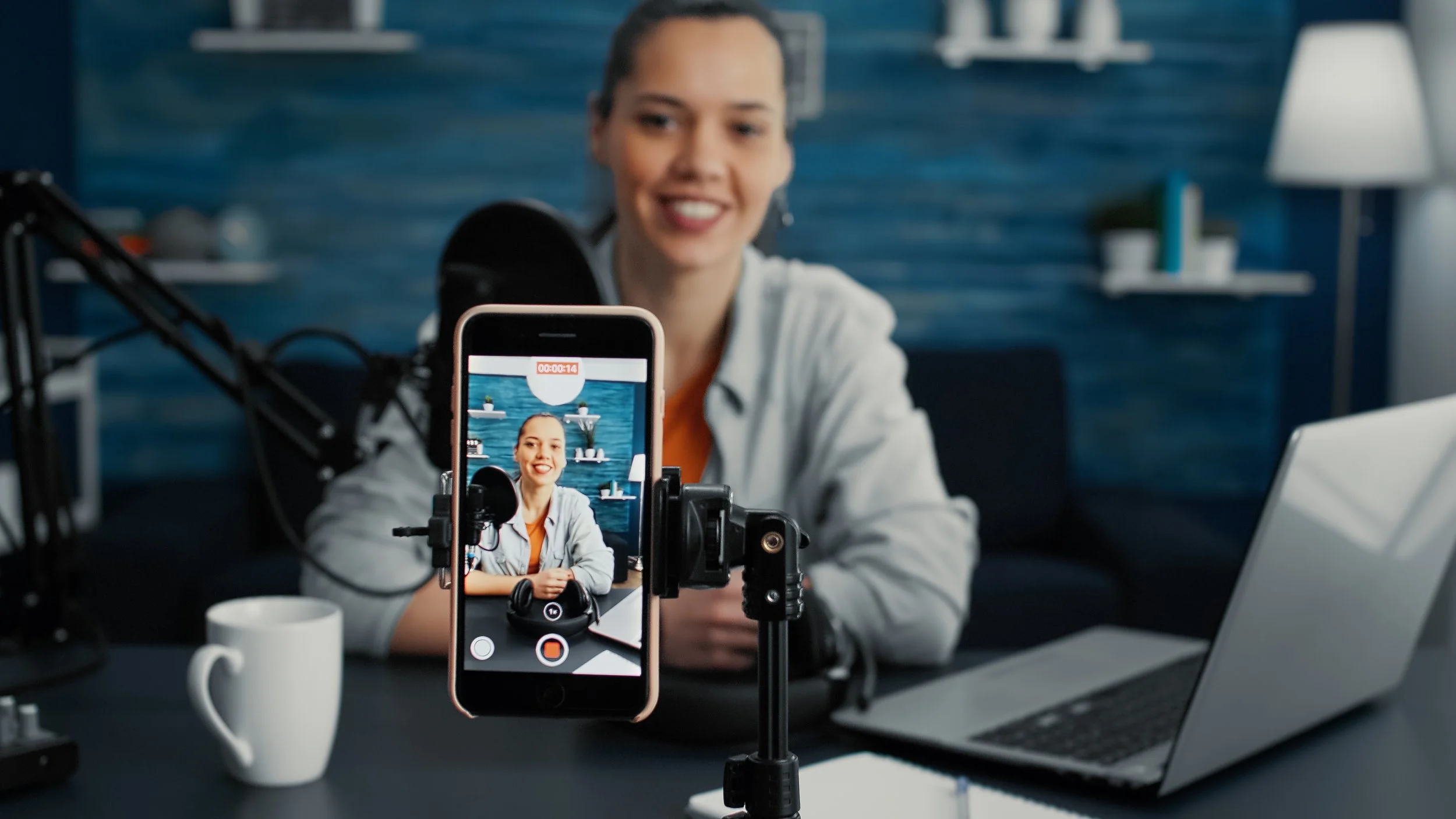 A woman recording a video with her smartphone in front of a microphone, with a laptop, a white coffee mug, and a blue wall with shelves in the background.