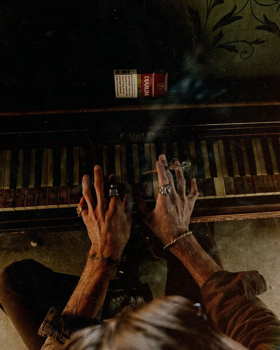 A person playing a vintage piano with hands adorned with rings, smoking a cigarette. The piano is labeled 'Chime Grand' and has a pack of Djarum cigarettes on top.