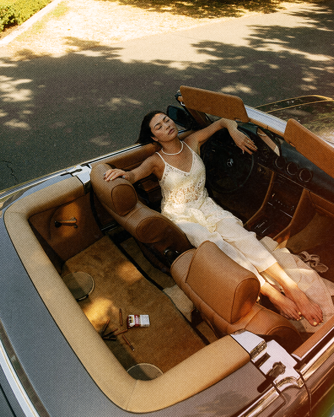 A woman in a white lace dress and pearl necklace relaxing in a vintage convertible car with tan interior on a sunny day.
