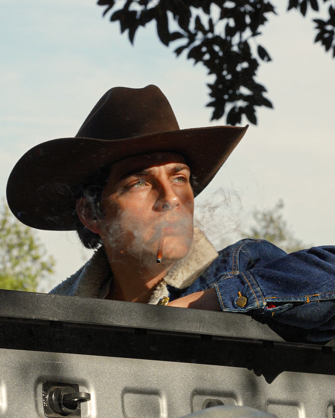 A man wearing a cowboy hat and denim jacket leaning on a vehicle, smoking a cigarette, with trees and a cloudy sky in the background.