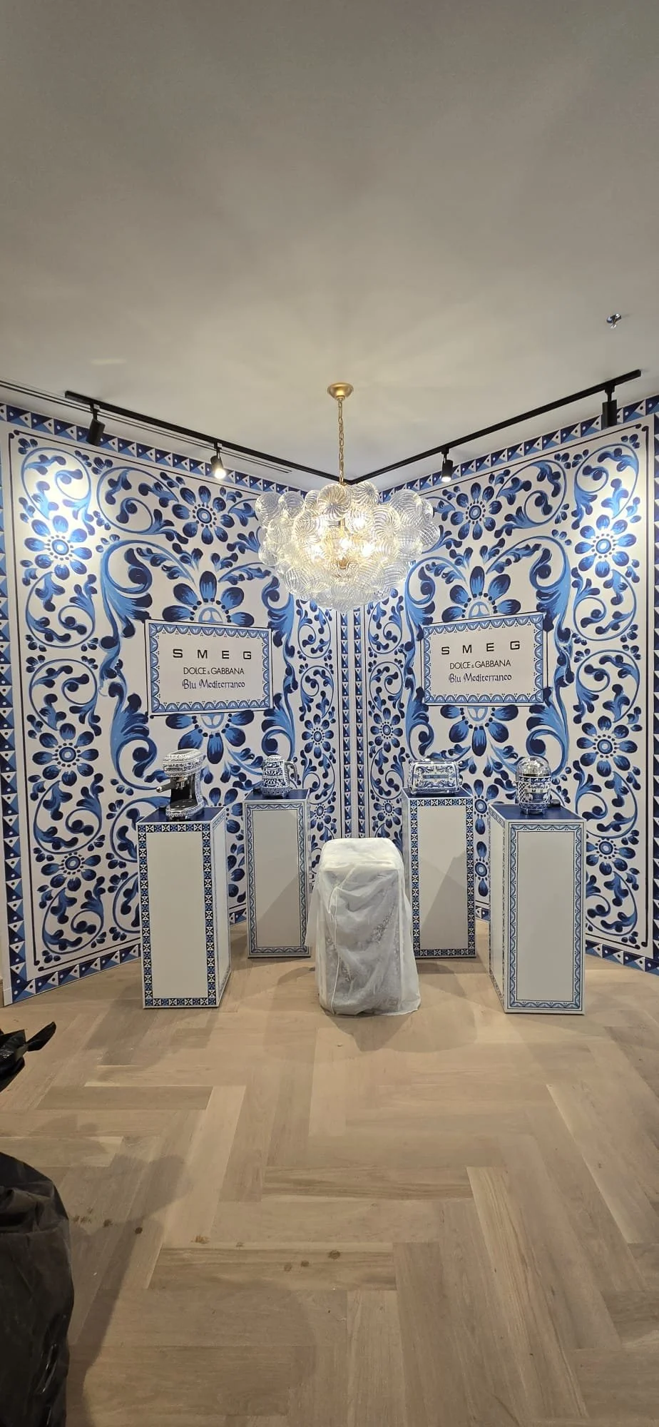 A display area with blue and white decorative patterned walls and three white pedestals showcasing handbags. A white cloth covers an object in the center, with a chandelier hanging from the ceiling.