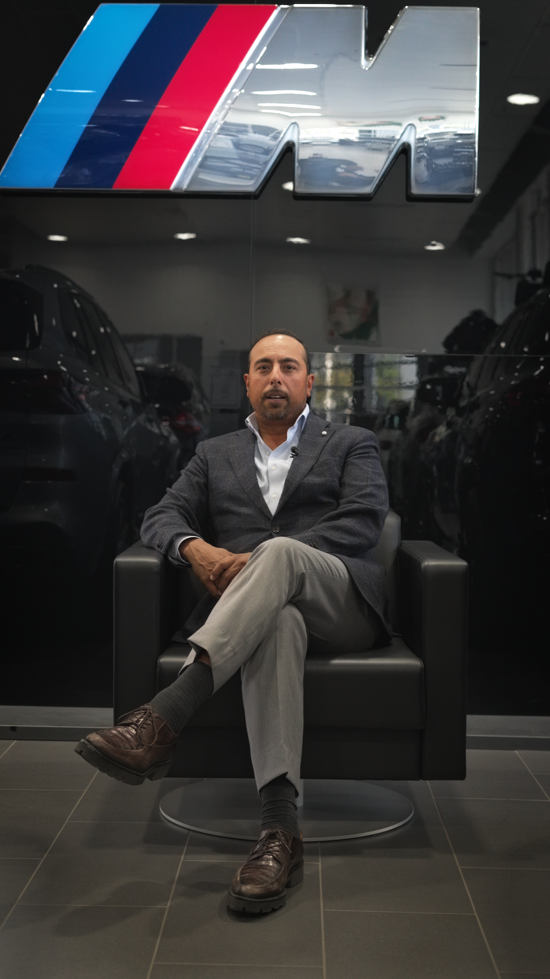 A man in a grey suit sitting on a black armchair in front of a black wall with a large BMW M logo.