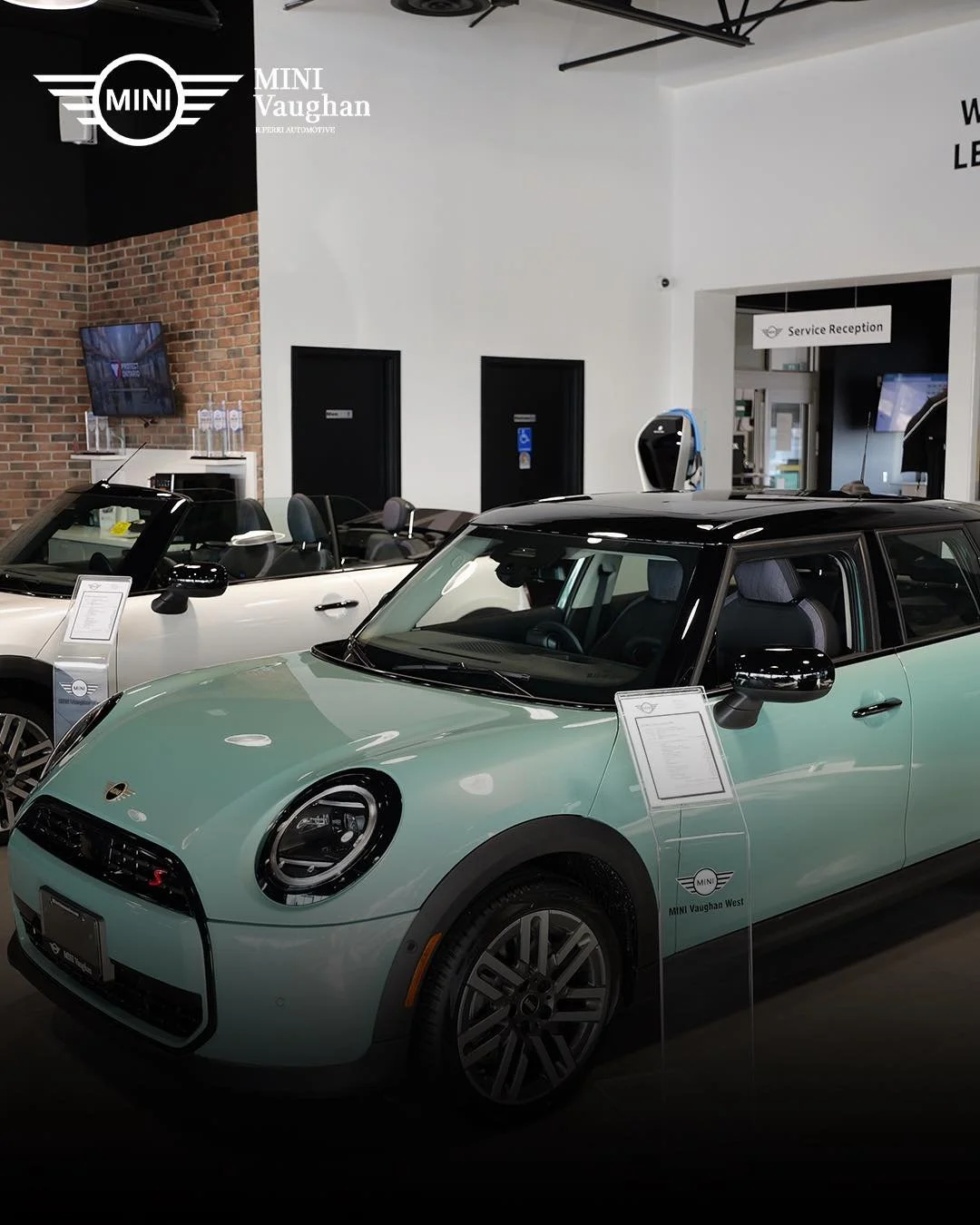 Display of a mint green Mini Cooper with a black roof in a car showroom, with a white convertible Mini Cooper behind it and a service reception area visible in the background.