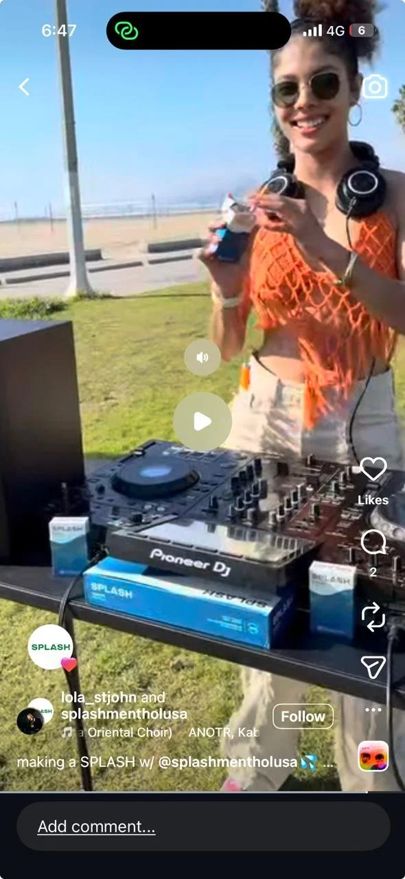 A woman with sunglasses and hoop earrings, wearing an orange mesh top over a beige outfit, stands outdoors at a DJ setup with Pioneer DJ equipment and a Splash brand box on a table. She is smiling and holding a phone and a small bottle.