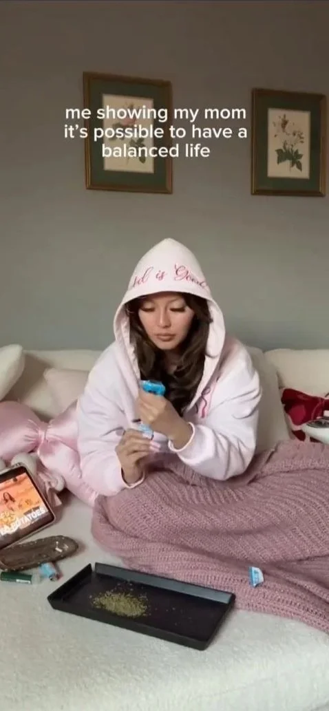 A young woman sitting on a bed, wearing a pink hoodie with the hood up, looking at her phone. The bed has a pink blanket, and there are various items on it, including a rolling tray with marijuana, a lighter, and a smartphone. The wall behind her has two framed pictures, and the text overlay says, "me showing my mom it's possible to have a balanced life."