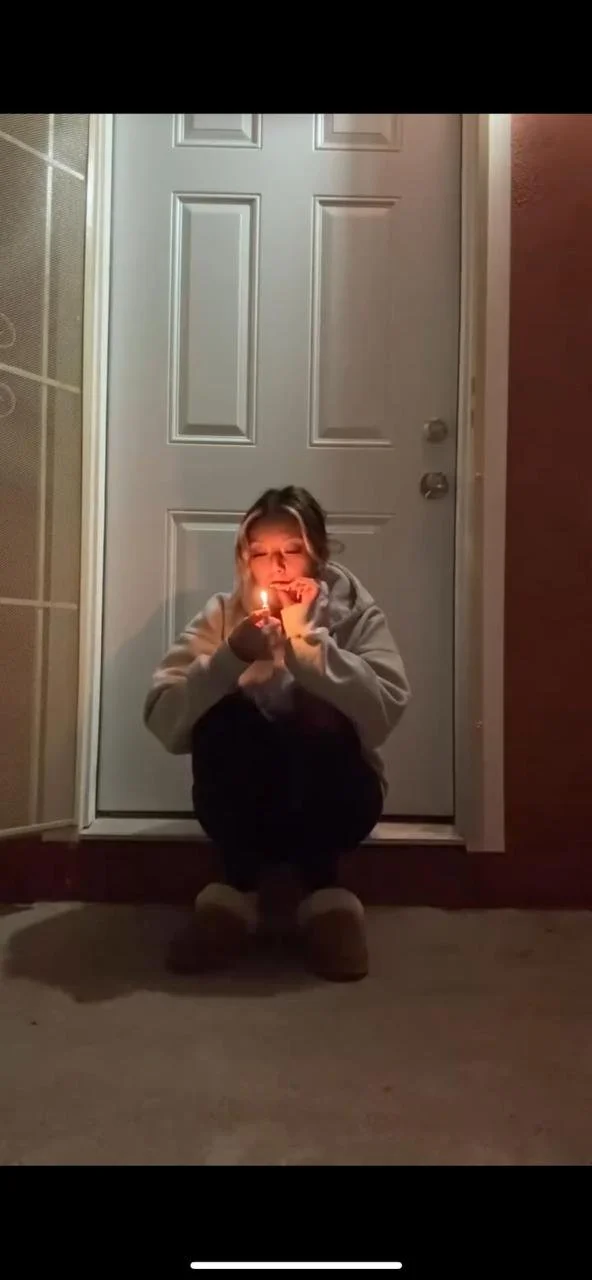 A young girl is crouching outside a door at night, holding a lit candle close to her face, creating a warm glow.