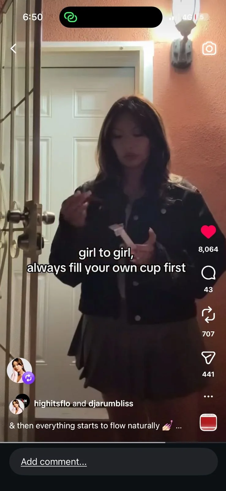 A woman standing at a door with a light-colored door behind her. She has shoulder-length dark hair and is wearing a dark jacket and a skirt. She is holding a packet and appears to be applying lipstick. The scene is illuminated by an exterior wall-mounted light fixture.