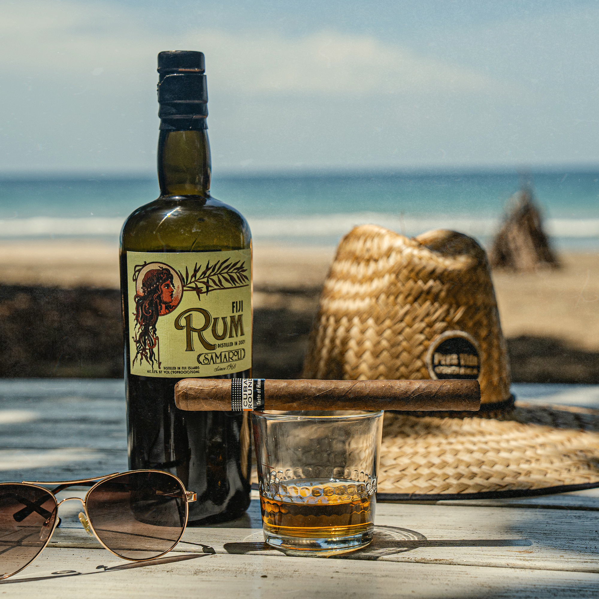 A bottle of rum, a glass with a drink, a cigar, sunglasses, a straw hat, and a beach in the background.