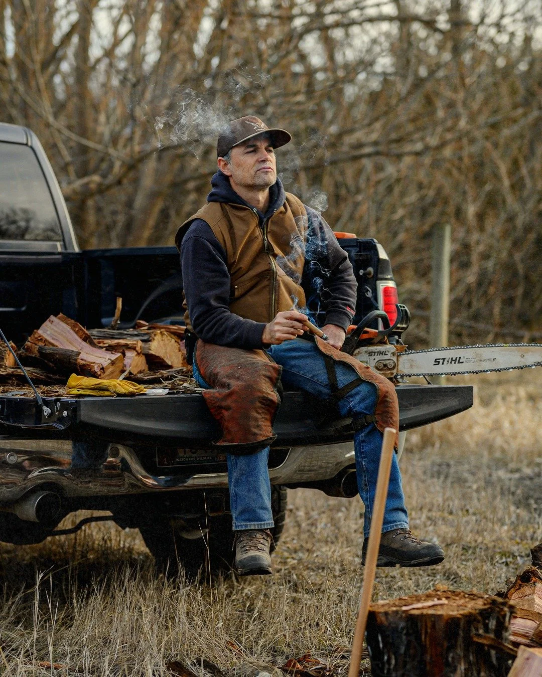 A man in work clothes sitting on the tailgate of a black pickup truck, smoking a cigar, with chopped wood and a chainsaw nearby, outdoors in a wooded area.