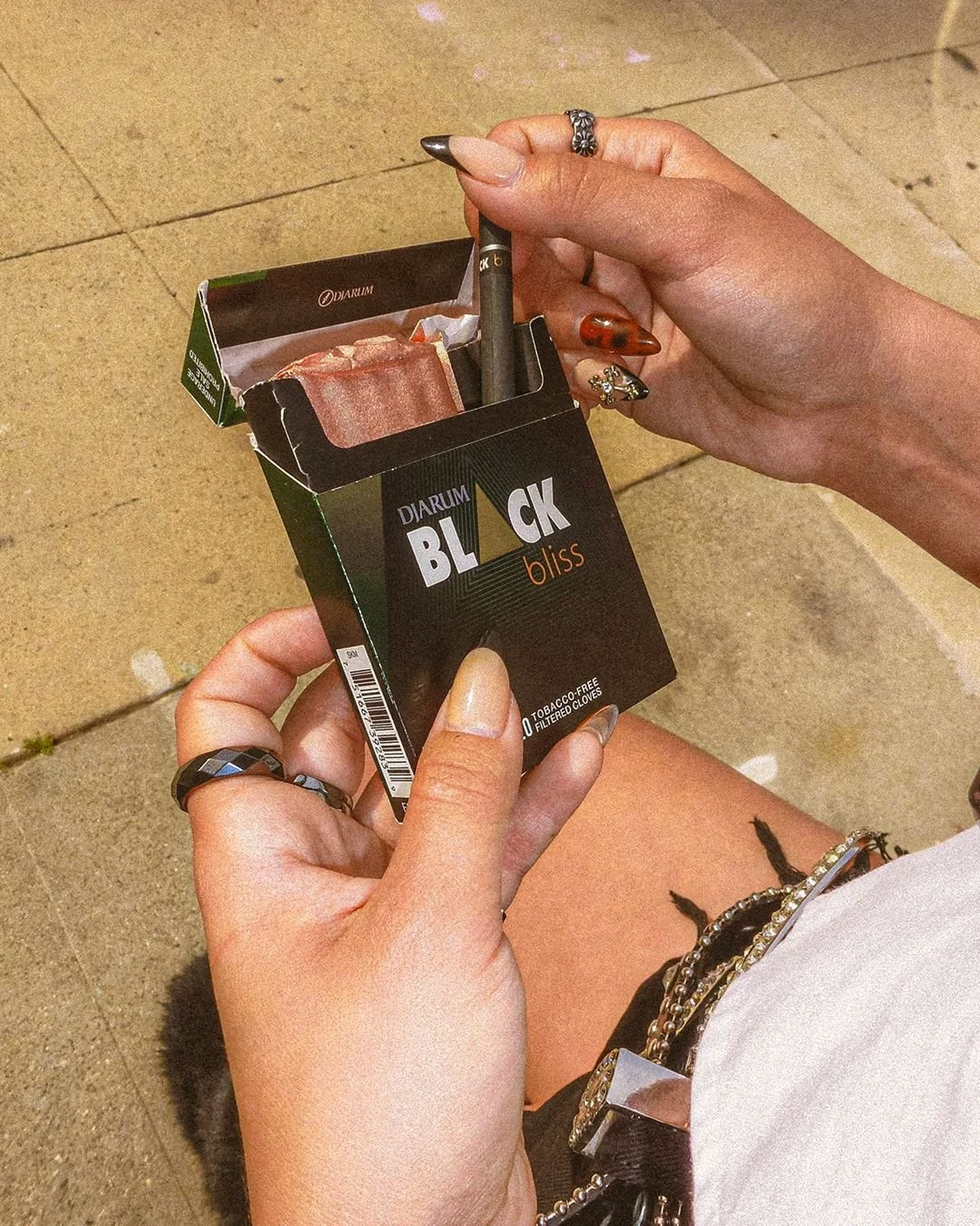 A person holding a box of Black Bliss tobacco-free, filtered cigarettes, with a vape pen sticking out from the opening of the box, on a tiled floor, wearing multiple rings and bracelets.