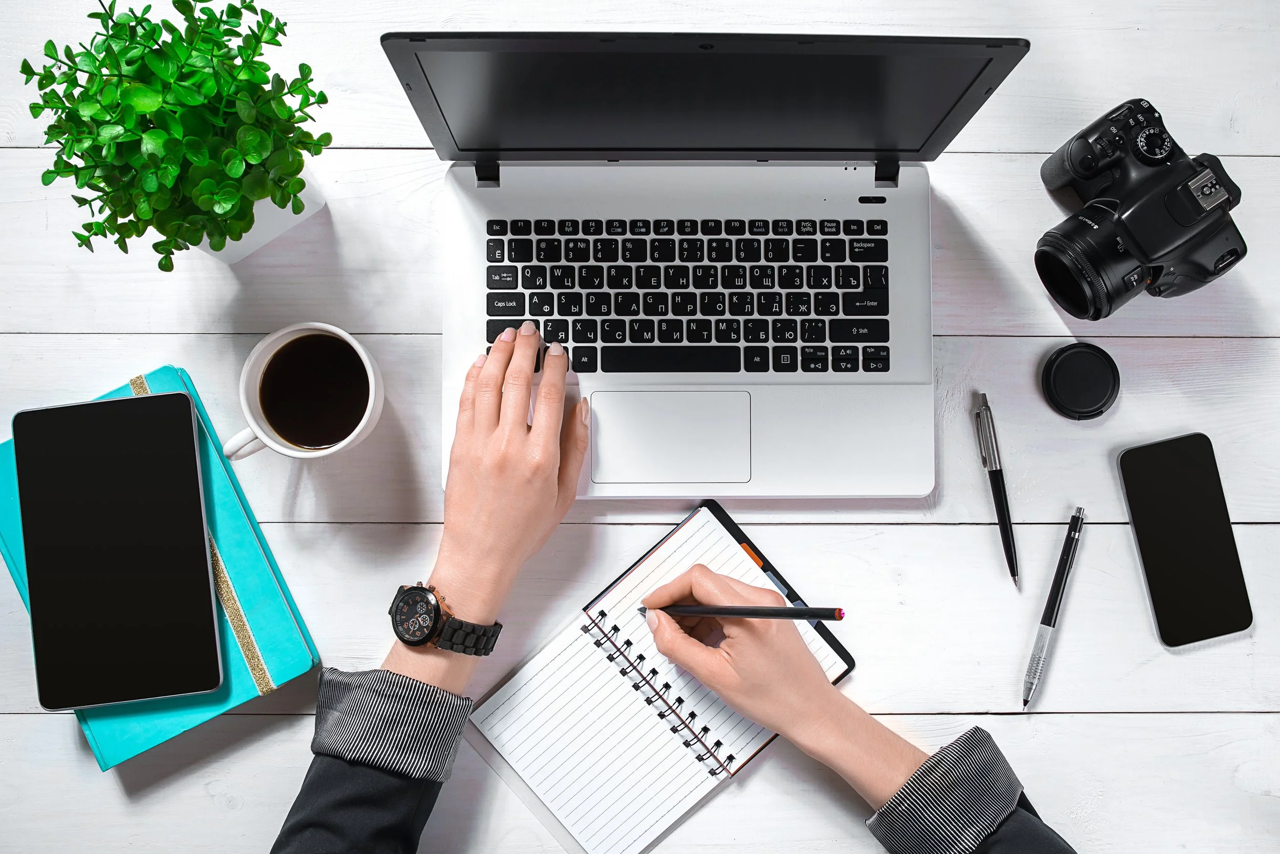 Overhead view of person working at a white desk surrounded by a laptop, camera, phone, notebooks, pens, coffee, and a plant.