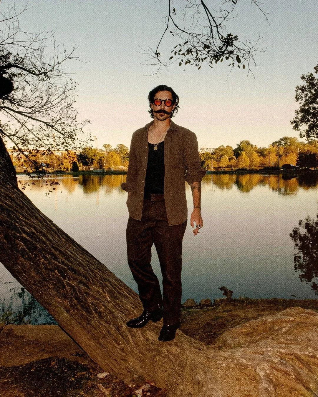 Man with dark wavy hair, sunglasses, and a mustache, standing on a bent tree trunk by a lake at sunset, wearing a brown shirt, black T-shirt, dark pants, black shoes, and some jewelry.