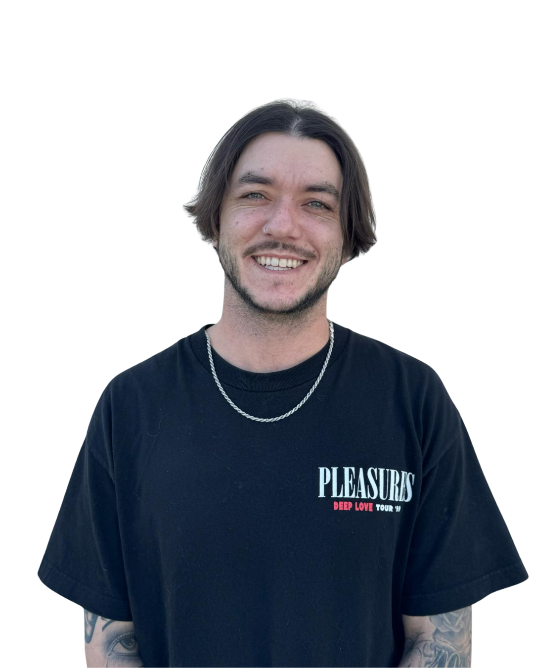 A young man with medium length brown hair, smiling, wearing a black t-shirt with the word 'PLEASURES' printed on it and a silver chain necklace.