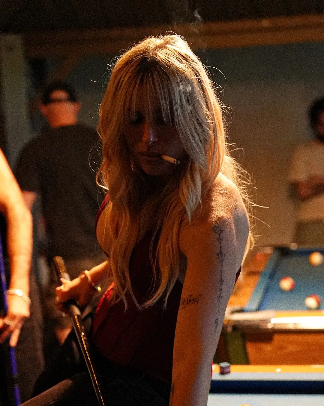 Woman with long blonde hair smoking a cigarette and holding a pool cue at a dimly lit bar or pool hall.