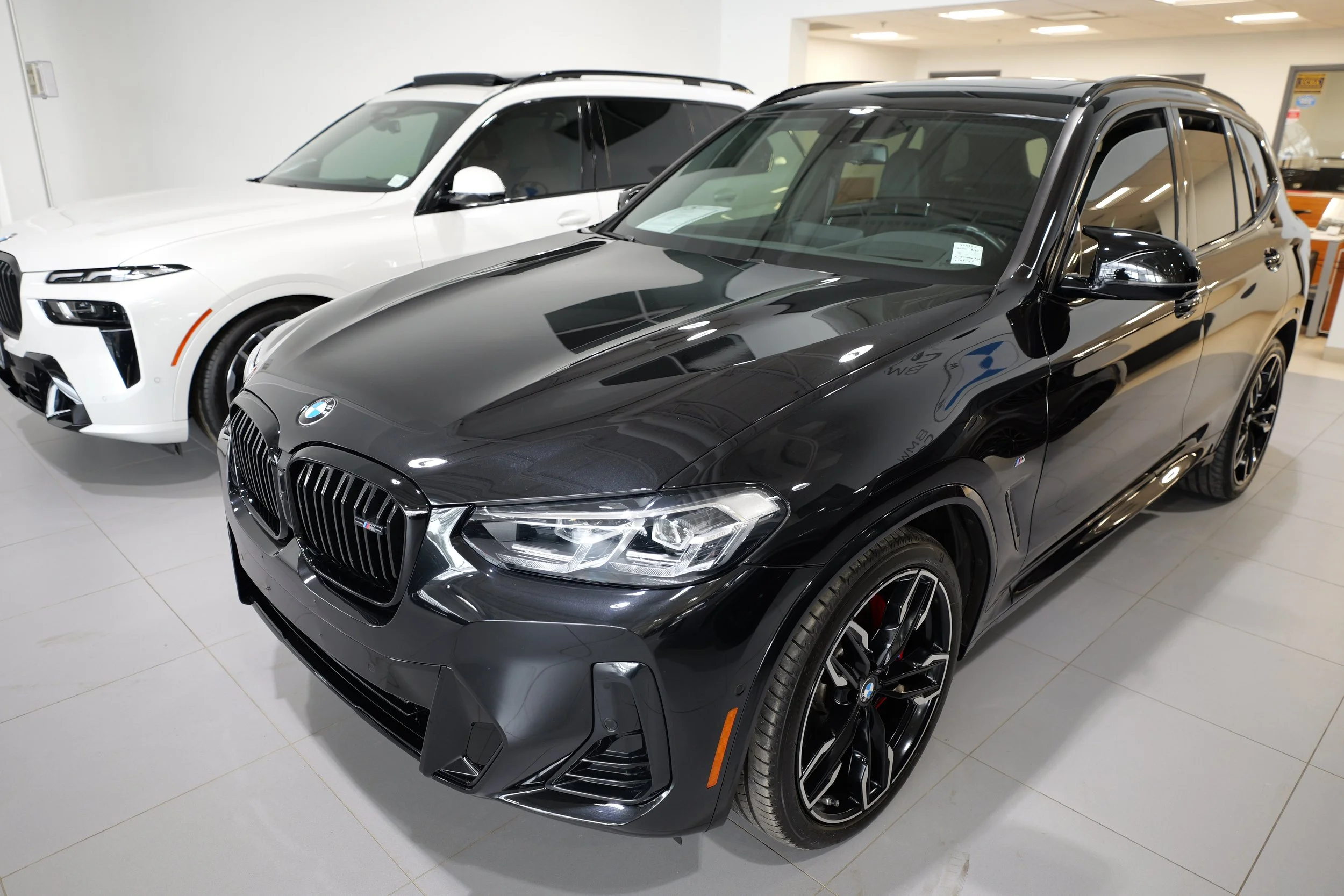 A black BMW SUV parked indoors next to a white luxury SUV, both inside a well-lit car dealership.