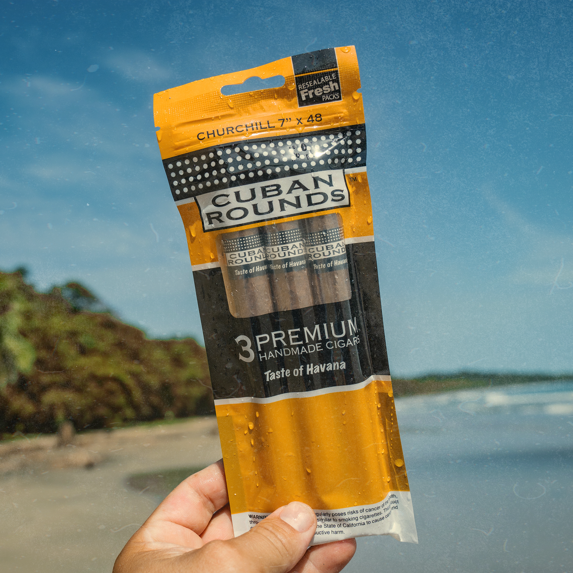 A hand holding a package of Cuban Rounds cigars with a beach and ocean in the background.