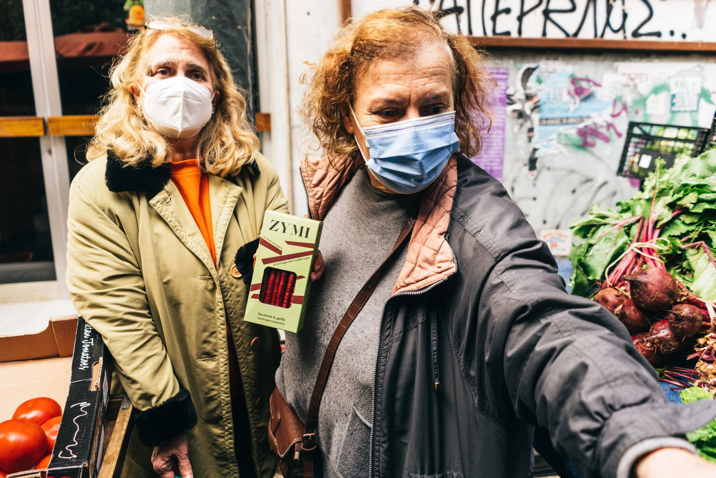Two people wearing face masks hold a box of ZYMI outdoors in a farmers' market with colorful graffiti visible in the background.