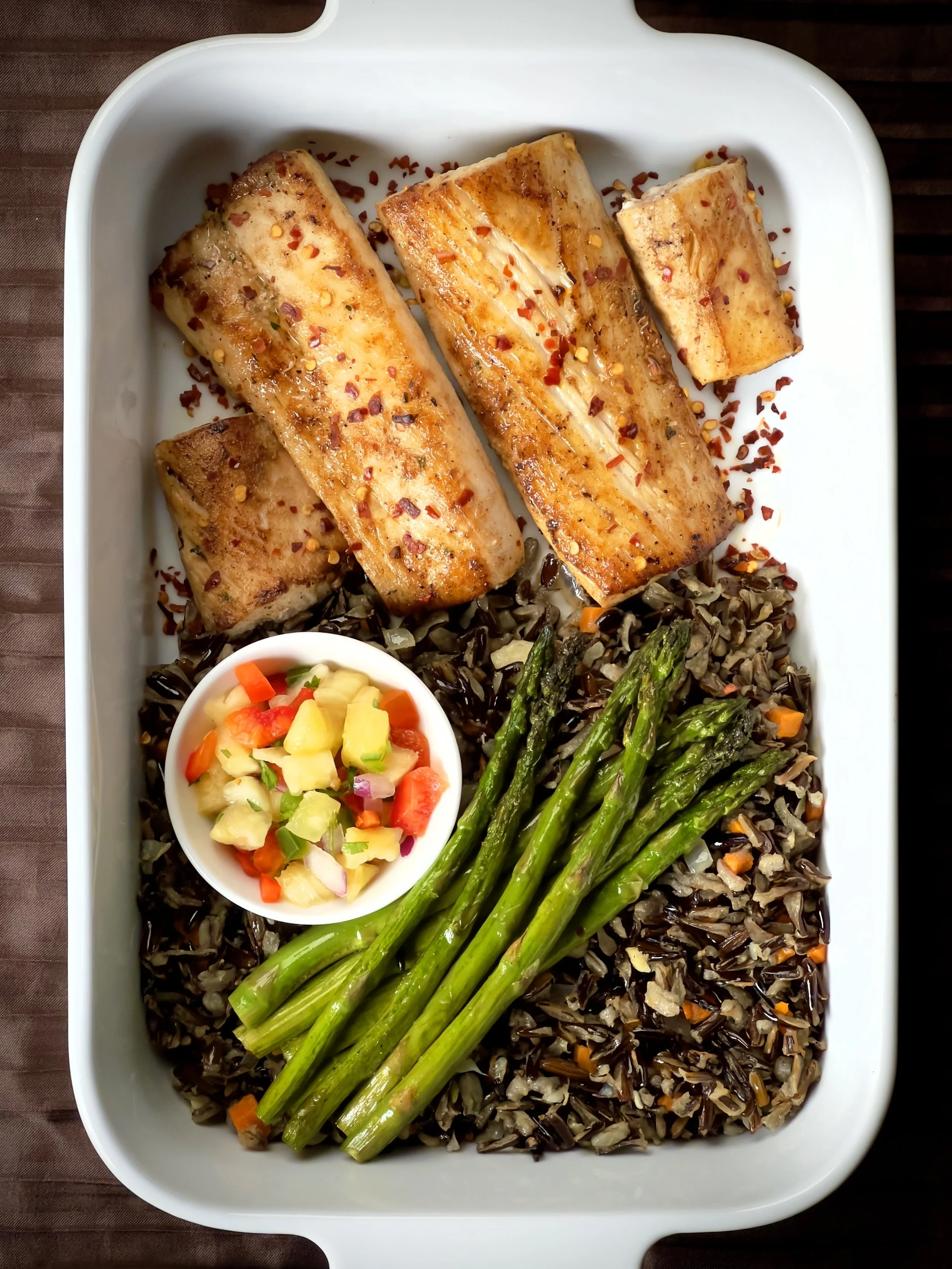 Baked salmon fillets topped with red pepper flakes, served with black rice, grilled asparagus, and a small bowl of pineapple salsa, on a white rectangular platter.