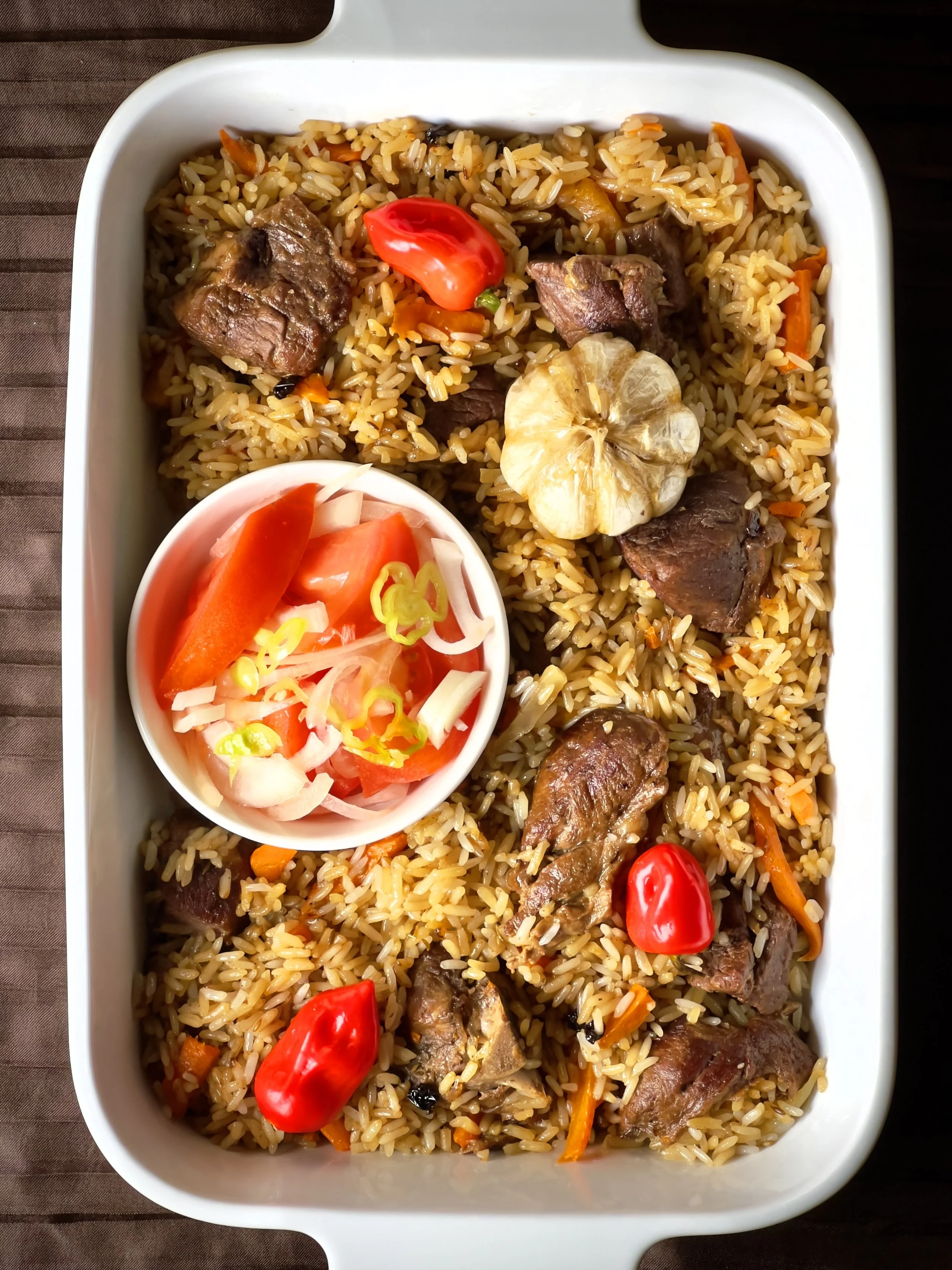 A white rectangular dish containing cooked rice with beef, carrots, and garlic, and a small bowl of sliced tomatoes and onions with green chilies.