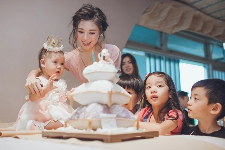 Young woman and children celebrating a birthday with a cake, a baby girl wearing a crown, and smiling kids around the table.