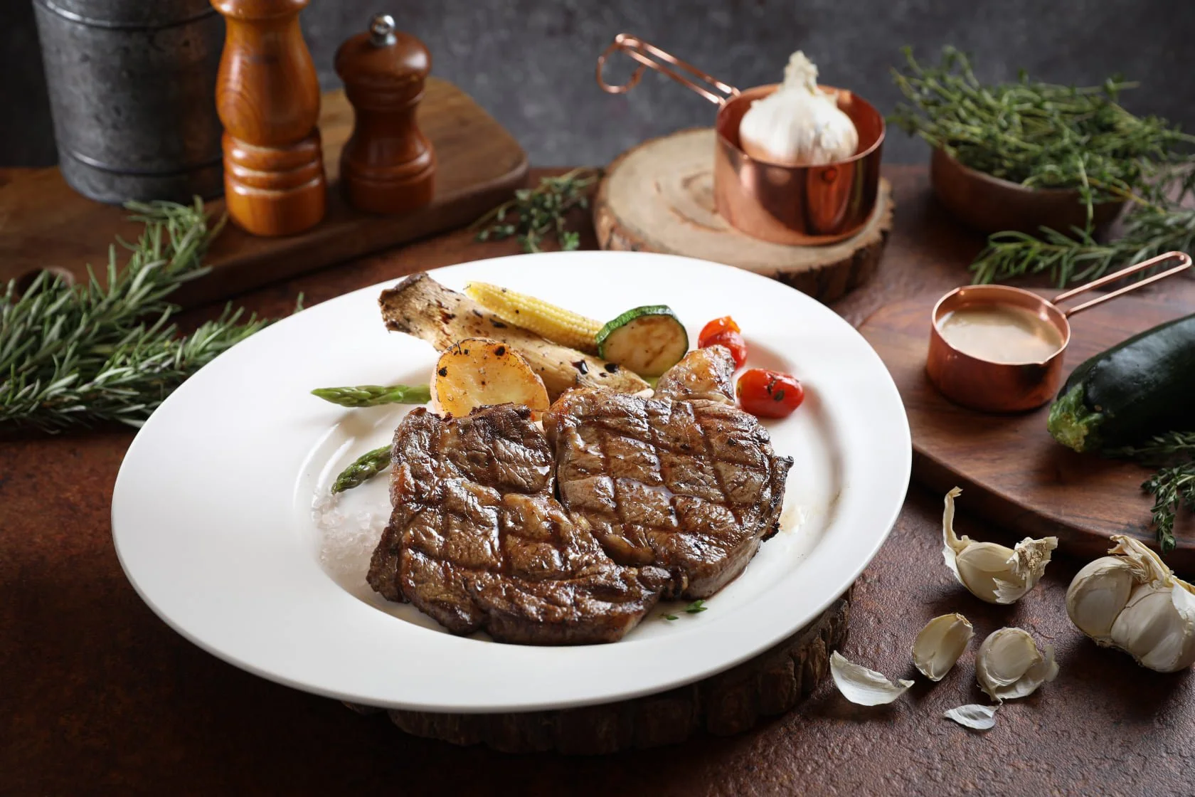A plate with grilled steak, grilled vegetables, and potatoes on a rustic table with herbs and seasonings in the background.