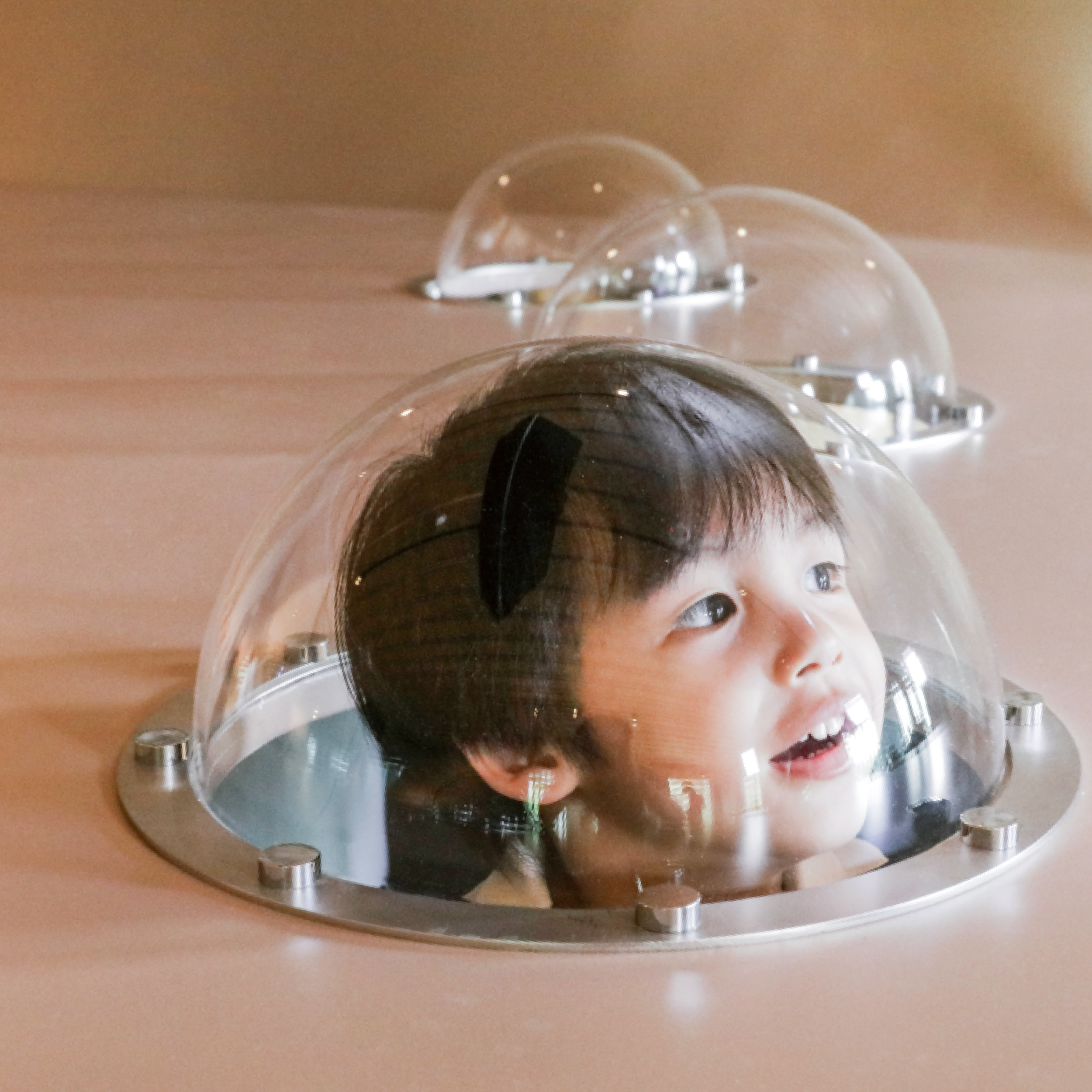 A young boy looking up through a transparent dome on a table, with a curious and happy expression.
