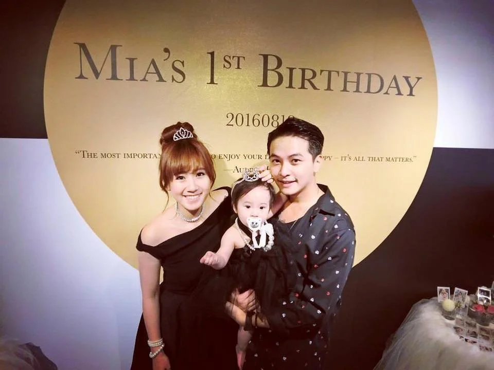 A family celebrating Mia's first birthday, with a woman, a man, and a baby girl dressed in black, all smiling, in front of a large gold and black backdrop with birthday details.