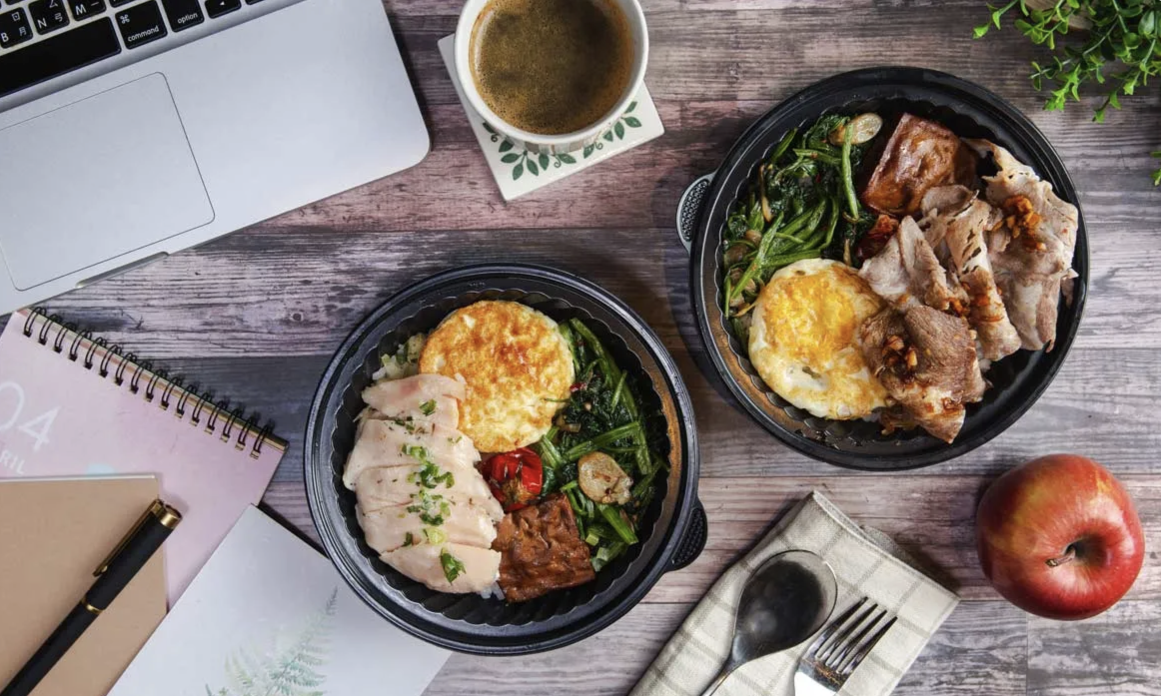 Two takeout containers with food, a cup of coffee, an apple, a spiral notebook, a pen, a fork, a spoon, and a napkin on a wooden table.
