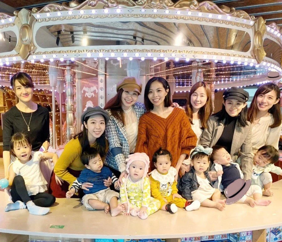 A group of women and young children posing together in front of a carousel ride at an amusement park. The women are smiling, and the children are sitting or standing on a platform, some holding toys or wearing hats.