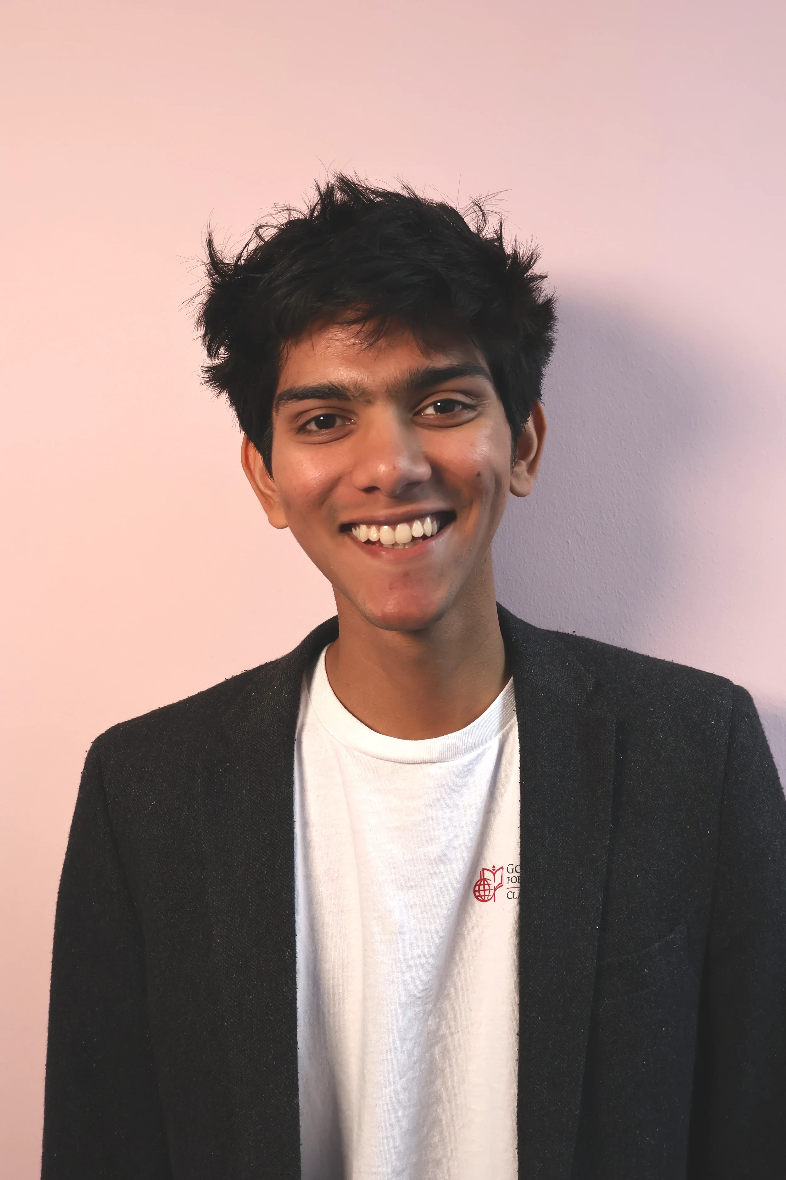 Portrait of a person with curly hair and a black scarf, wearing a dark jacket, set against a light brown background.