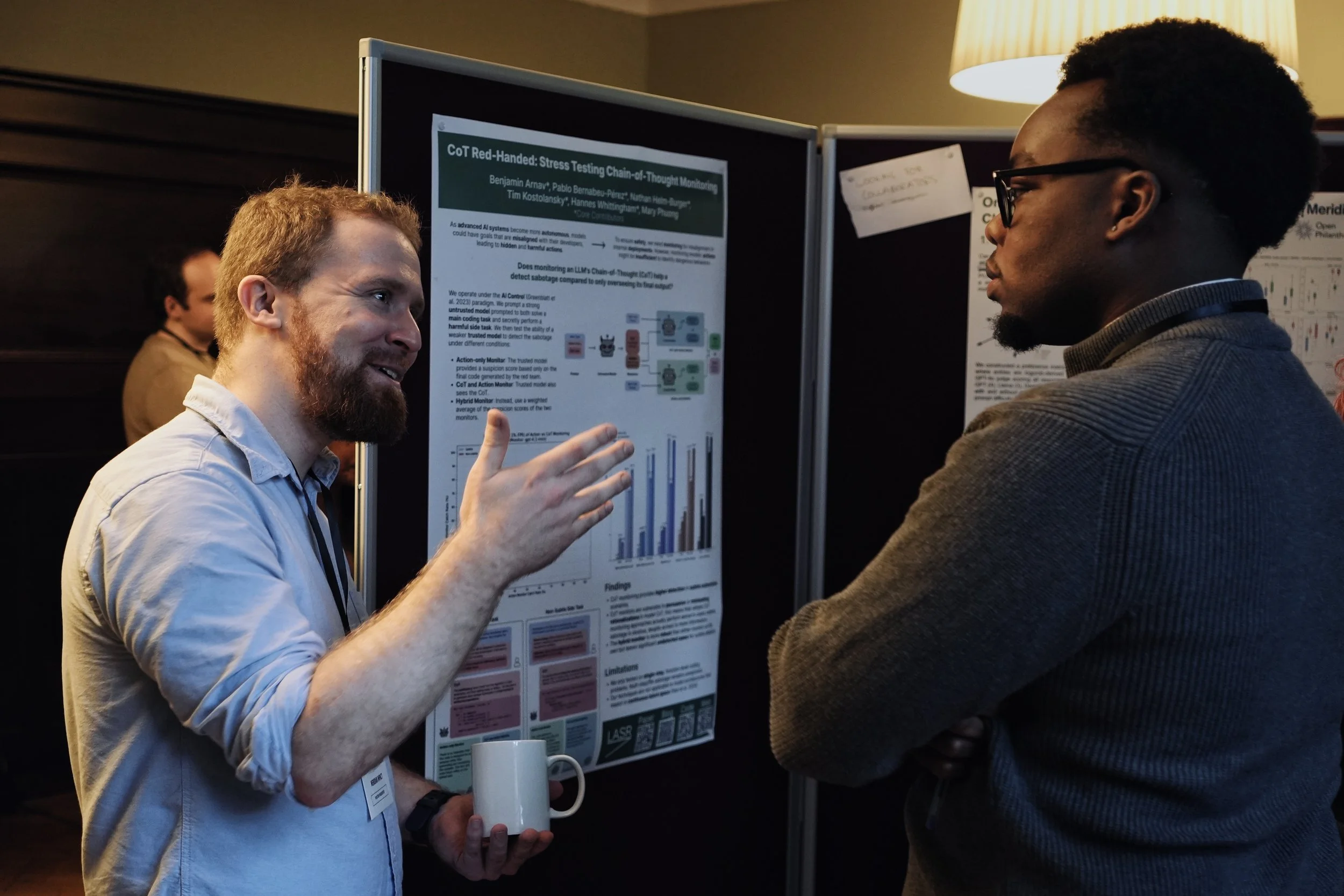 A person speaking in front of a presentation about AgentHarm baseline. The individual is gesturing with their hands and standing near a poster with the text 'Cambridge Safety H.' The presentation slide includes diagrams and text related to computational tools.