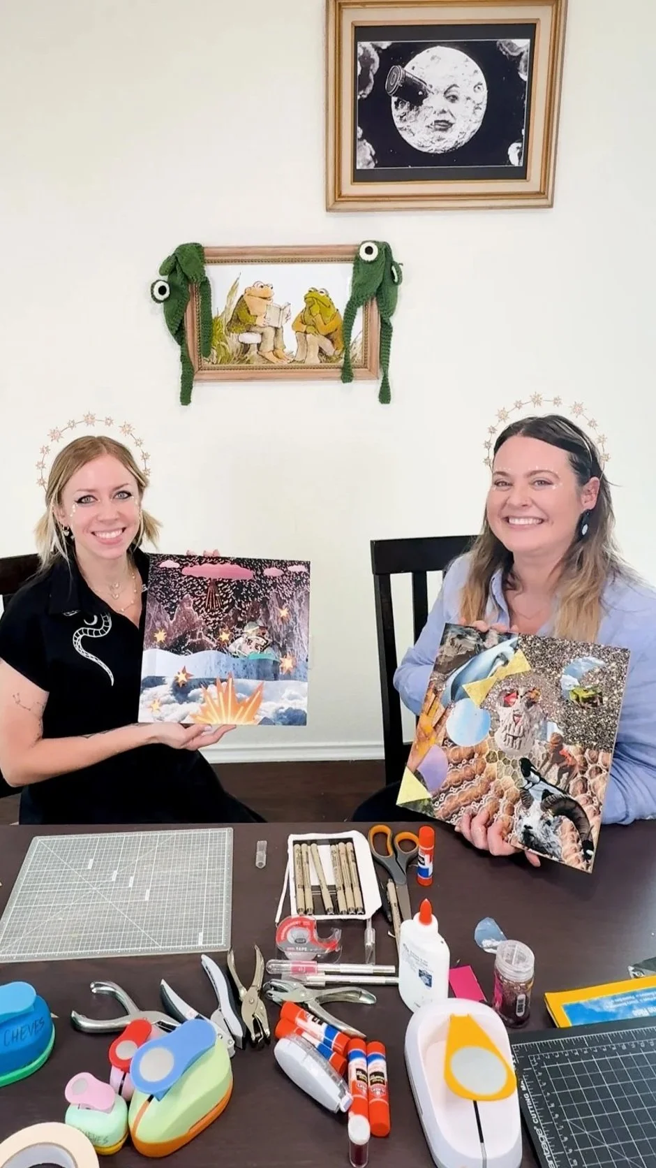 Two women sit at a table, each holding a colorful, collage-style artwork. The table is covered with craft supplies like glue, scissors, tape, and cutting tools. In the background, two framed puppet-like frog characters sit on a wall, and above them is a framed black-and-white moon illustration with a face.