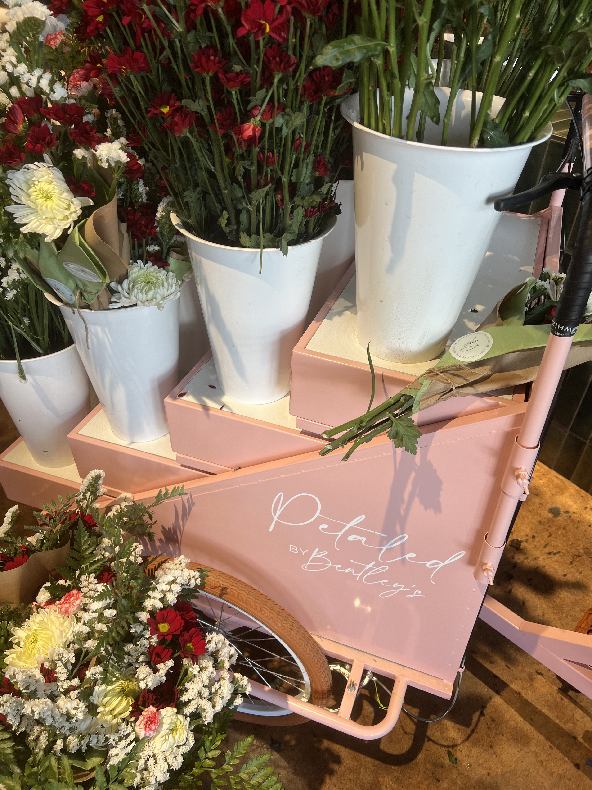 A pink floral cart with white potted flowers and green plants, labeled "Petaled by Bentley's," with a bouquet of mixed flowers in front.