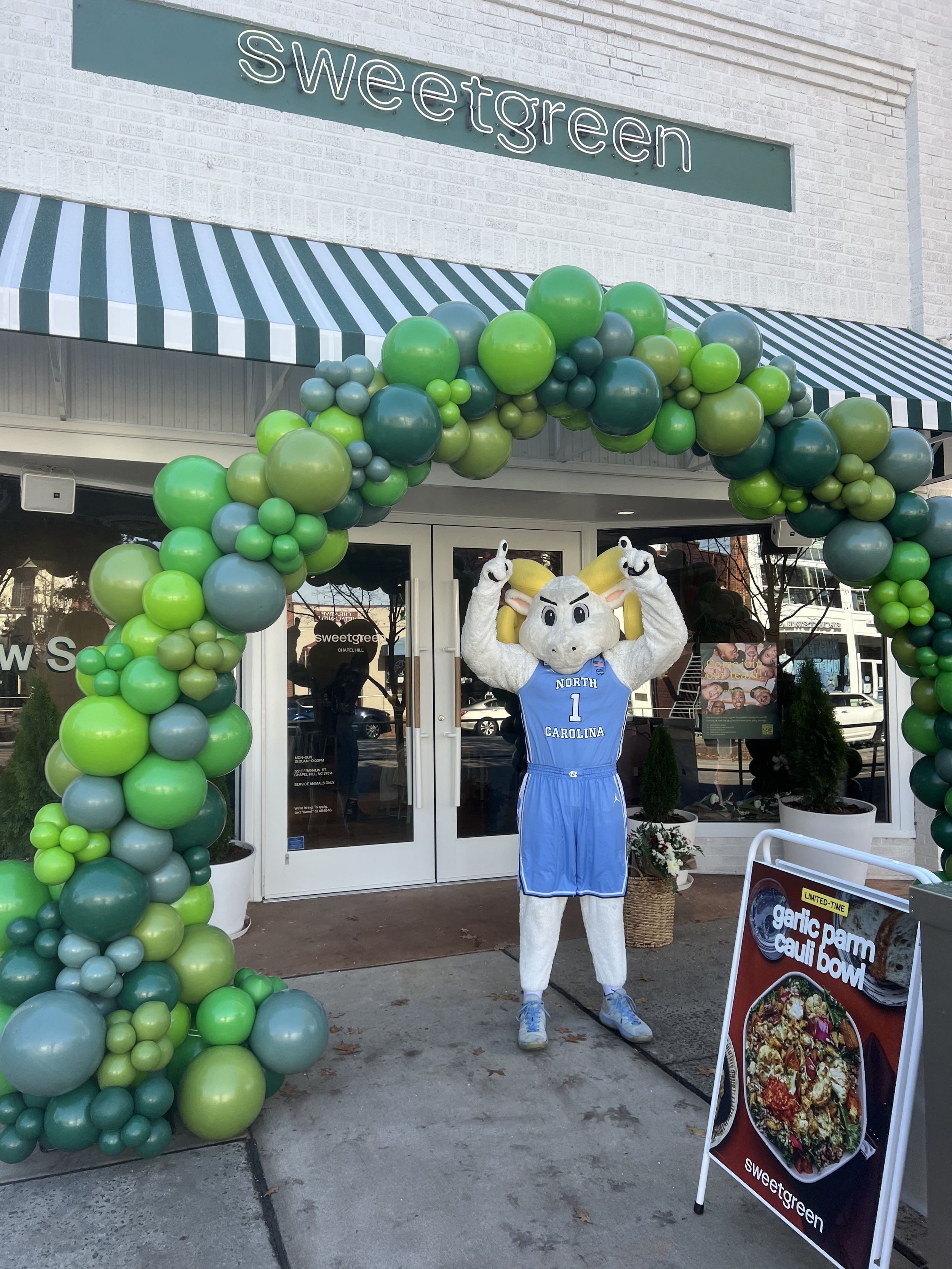 A mascot dressed in a North Carolina basketball uniform standing under a green balloon arch outside a storefront with a sign that says 'sweetgreen'.
