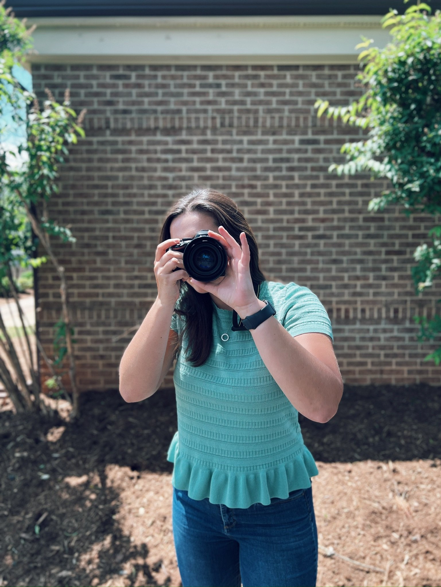 A woman taking a photo with a camera outdoors, standing in front of a brick wall and greenery.