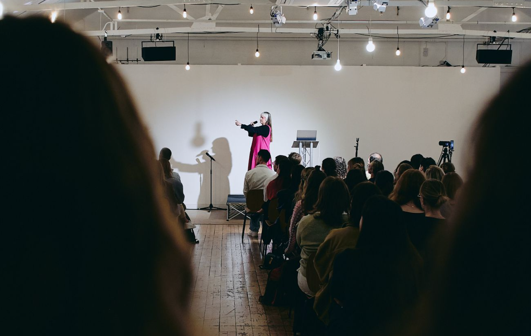 A woman with blonde hair in a colorful jacket and red striped shirt pointing at a projected presentation on a wall.