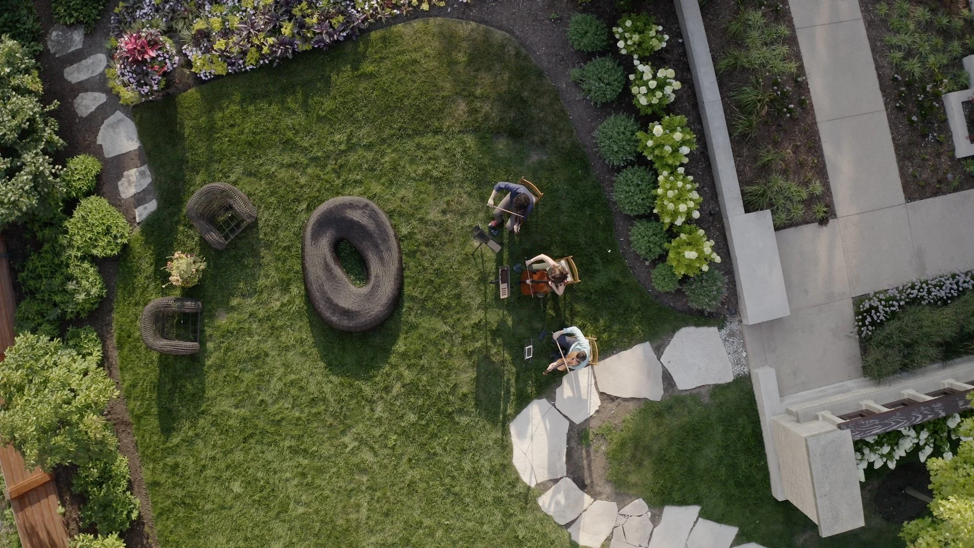Overhead view of a backyard garden with a lawn, flower beds, stone walkway, and three people playing string instruments, possibly a small ensemble, seated on chairs along with music stands.