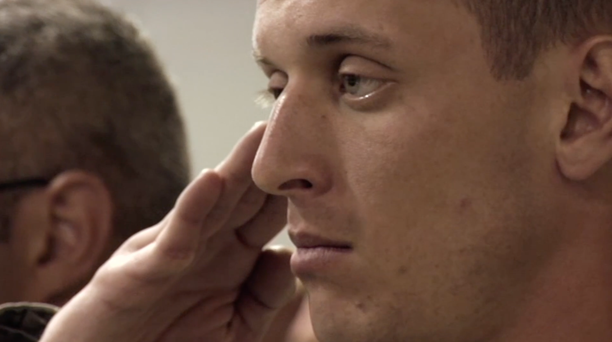 A close-up of a young man with light skin and short hair, looking contemplative, with someone touching his face.